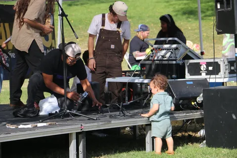 A DJ works at a turntable setup while a toddler in overalls stands nearby.
