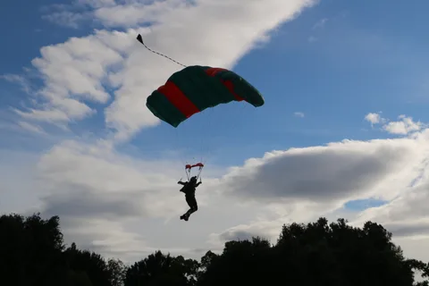 A skydiver descends under a teal and red parachute against a blue sky with clouds.
