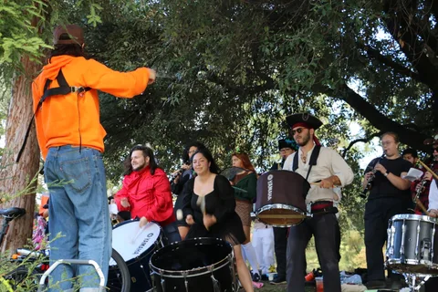 A band leader in an orange hoodie conducts drummers and horn players from atop a step stool.