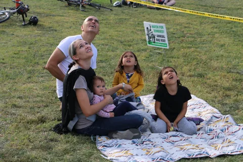 A family of five looks up in wonder at the skydivers, a Watch For Zombies bike ride sign beside them.