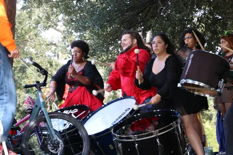 Drummers laugh mid-performance, drumsticks raised, with bikes parked beside them.