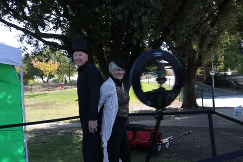 Two people, one in a dark cloak costume, stand near the photo booth light ring.