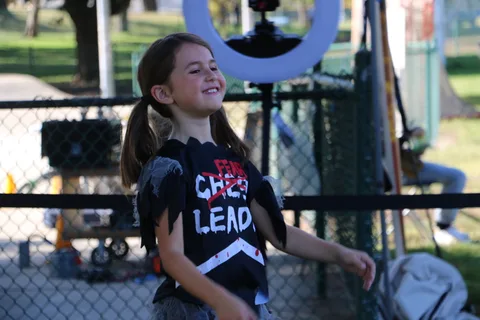 A girl in a cheerleader shirt smiles brightly near a photo booth ring light.