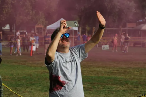 A man in a bloodstained shirt raises a hand and films the sky through drifting pink smoke.