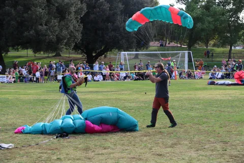 A landed skydiver gathers his parachute on the field as a crowd watches along the roped-off landing area.