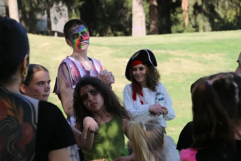 A person in colorful face paint stands with costumed friends including one in a pirate hat.