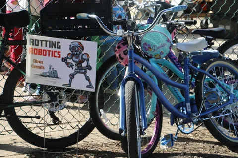A Rotting Robotics team sign from Citrus Circuits hangs on the fence beside parked kids bikes.