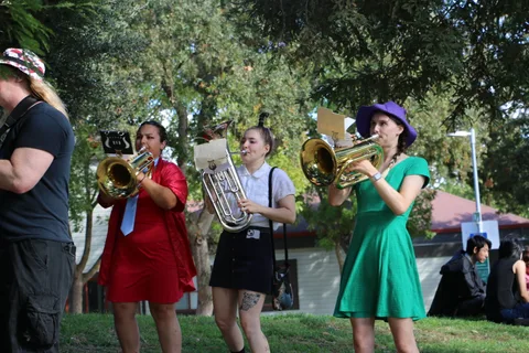 Three costumed brass players perform together, sunlight filtering through the trees.