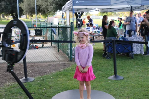 A girl in a pink skirt with an ear headband stands on the grass near event tents.
