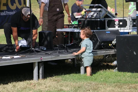 A small child walks near stage equipment as crew members set up sound gear.