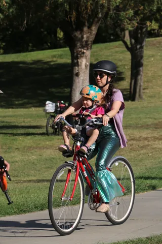 A mother in a mermaid costume rides with her child seated in front of her handlebars.