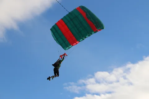 A skydiver under a green and red canopy fills the frame against an open sky.