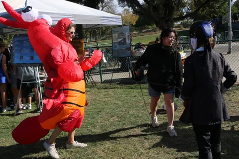 A woman in a giant inflatable lobster costume takes a photo while two fencers walk past.