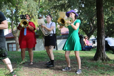 Three costumed brass players in colorful dresses perform under the trees.