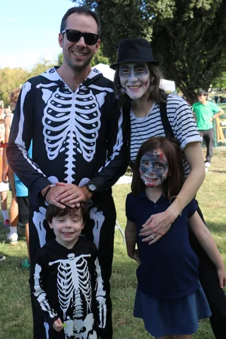 A family of four in skeleton costumes and zombie face paint poses together, the father covering his son's eyes playfully.