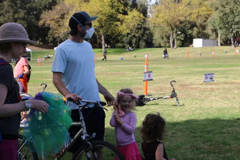 A man wheels a bike across the grass as a child with a green decoration stands nearby.