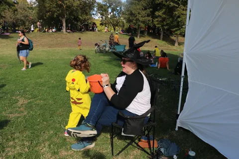 A woman in a witch hat holds hands with a child in a Pikachu costume on the grass.