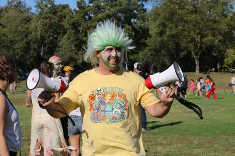 A laughing man in a green wig holds two megaphones, wearing an official Zombie Bike Parade shirt.