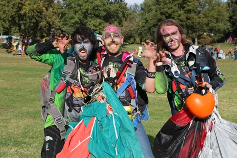 Three skydivers in zombie face paint growl and claw at the camera, parachutes bundled at their feet.