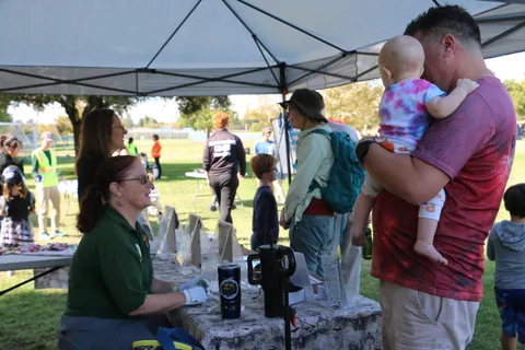 Families walk past event tents in the park, one woman carrying a baby.