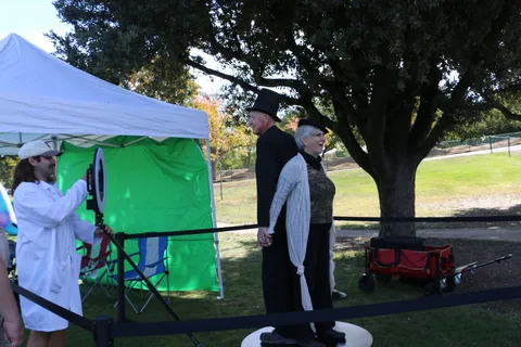Two people stand together near the photo booth equipment under the trees.