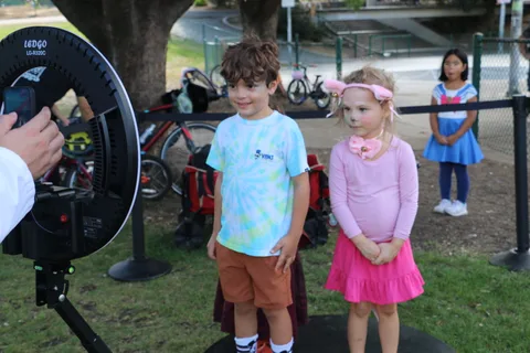 A boy and a girl stand together near a portable ring light in the park.
