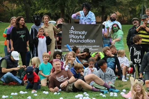 A costumed group including a clown and skeleton figures poses with children in front of a banner.
