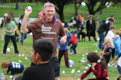 A man in an A Primally Playful Foundation shirt reaches up playfully as kids gather in the foreground.