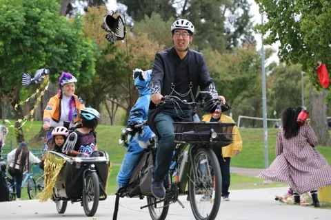 A man rides a cargo bike carrying children in a straw-decorated box as streamers fly overhead.
