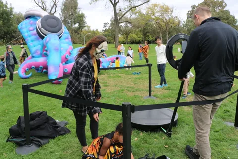 A woman in face paint stands near a camera rig with an inflatable figure in the background.