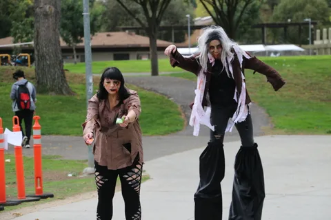 A zombie performer reaches toward the camera as a stilt walker looms behind.