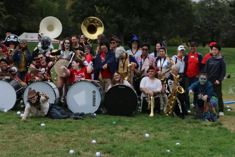 Band members holding sousaphones and drums gather for a group photo in the park.
