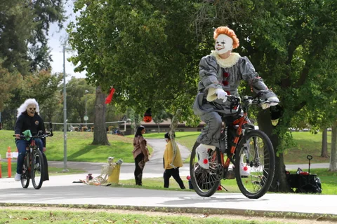 A person in a spooky clown costume with orange hair rides a bicycle along a park path.