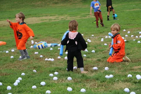 Two costumed children run across a grassy field scattered with white balls.