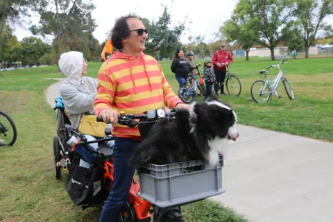A smiling man in a striped shirt rides with his dog seated in a front basket.