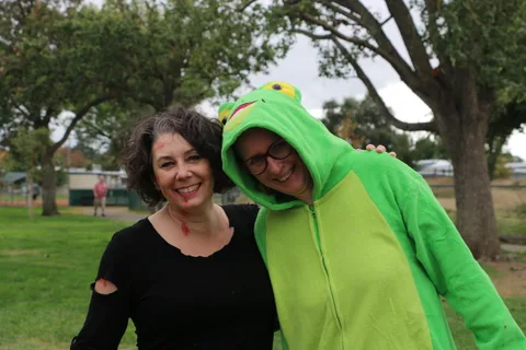 Two smiling women pose together outdoors, one wearing a green costume.
