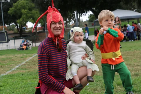 A man in a lobster costume holds a toddler dressed as a lamb beside a young boy.