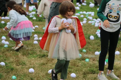 A child in a pastel dress and red cape holds white balls among other costumed kids on the grass.