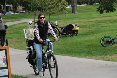 A woman in a skeleton-print jacket rides her bike along the park path.