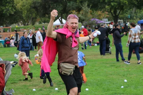A man in a pink cape raises his arm while holding a ball near a toddler.