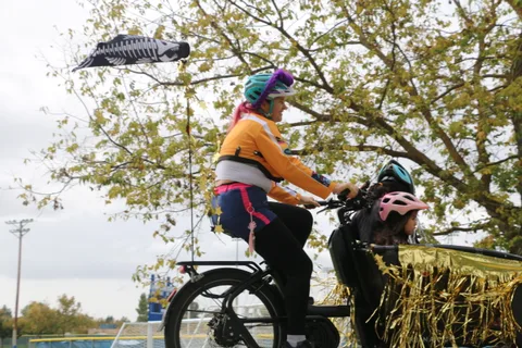 A cyclist in a colorful rainbow poncho rides with a child in a cargo attachment behind.