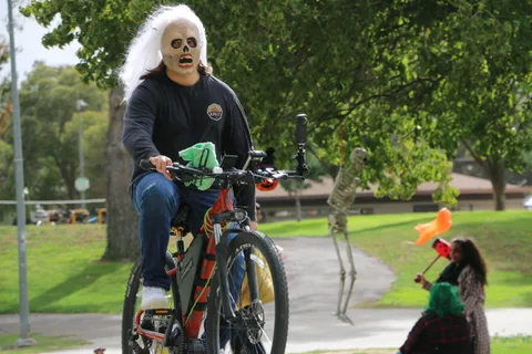 A woman in white ghostly face makeup rides a mountain bike past orange cones.