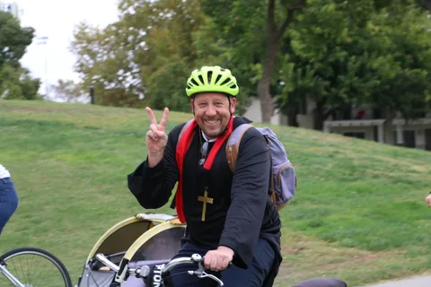 A man in priest-style costume flashes a peace sign while riding.