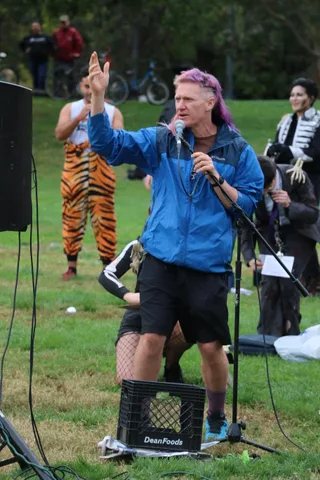 A man with pink hair speaks passionately into a microphone beside a tiger-costumed dancer.