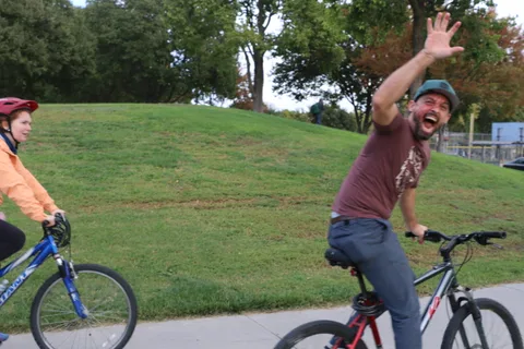 A man throws both arms up and grins widely while waving straight at the camera.