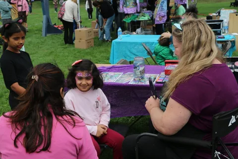Two girls sit together near a face painting booth, one already painted.