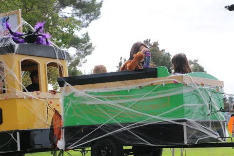 Children ride in a cobweb-decorated parade cart driven along the park path.