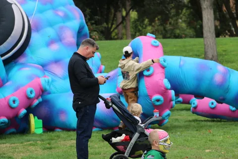 A man checks his phone near a stroller as a child touches a giant inflatable sculpture.