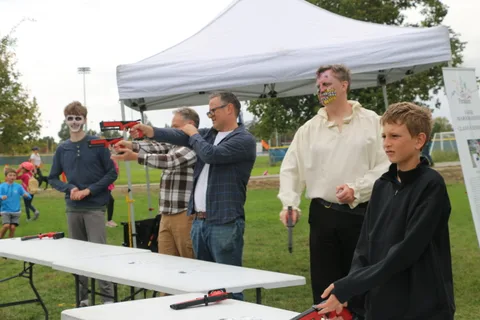 A group of men aim toy blasters at each other around a table under a tent.