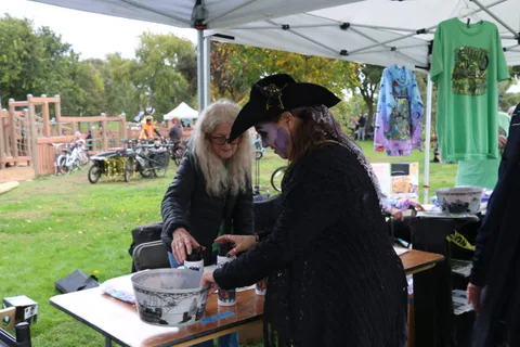 Two women in costume prepare items at a table under a tent.